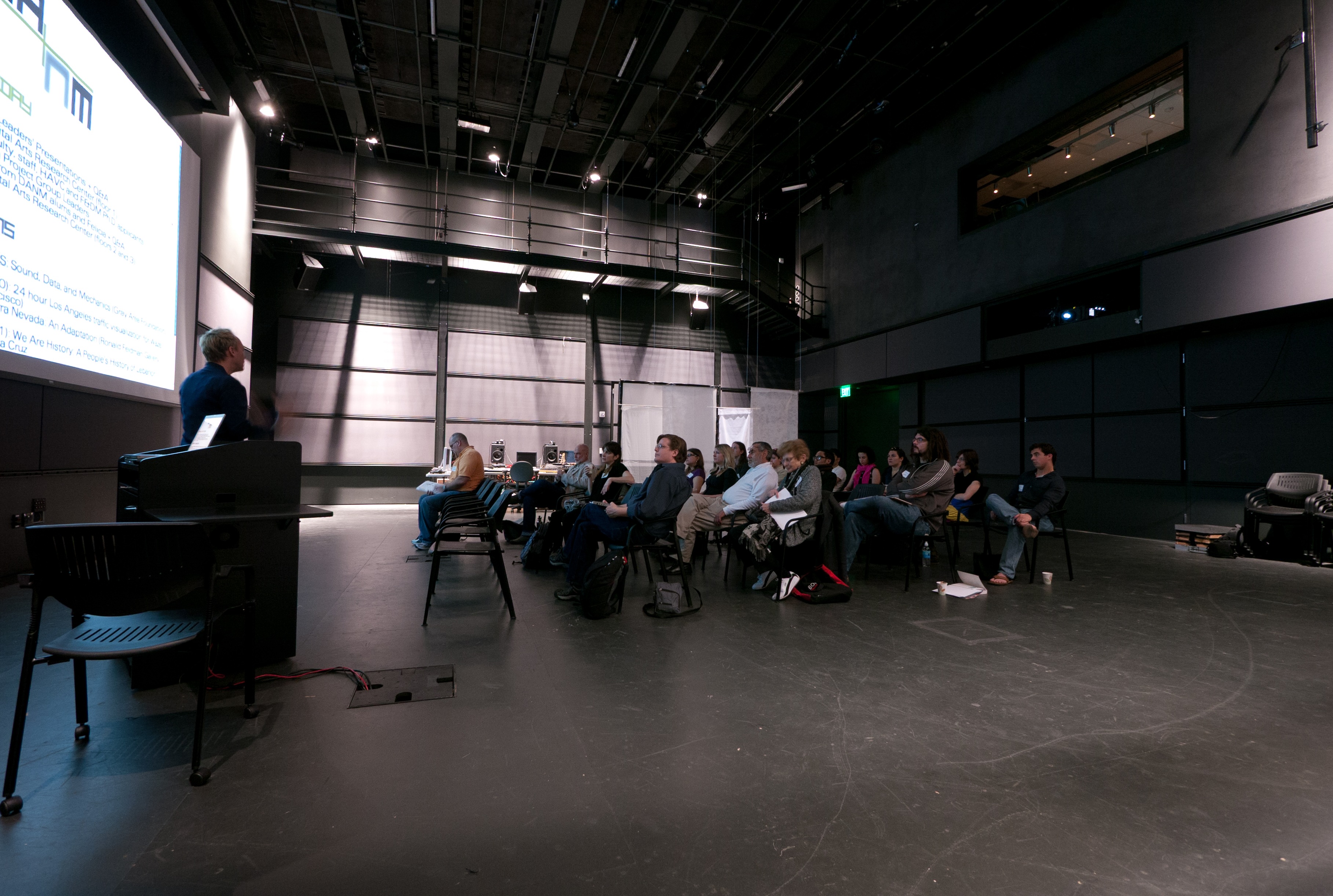 Faculty and Students sit in a dark big room, one man stands at the lectern, with a large white projection behind him of an agenda for the meeting.