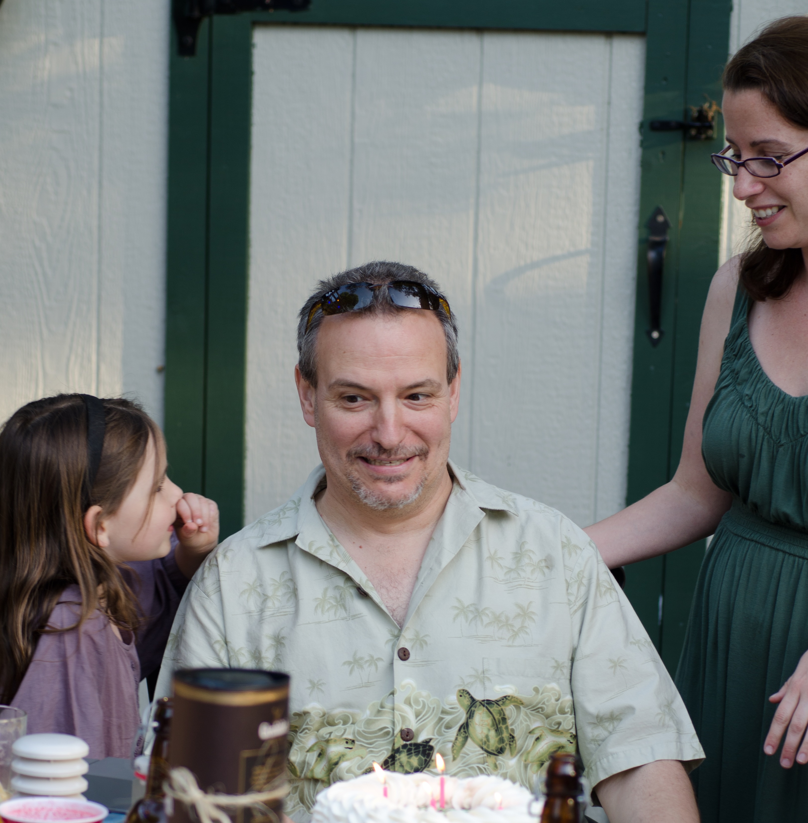 A man sitting at a table with a birthday cake in front of him. A girl on the left looking at the man.