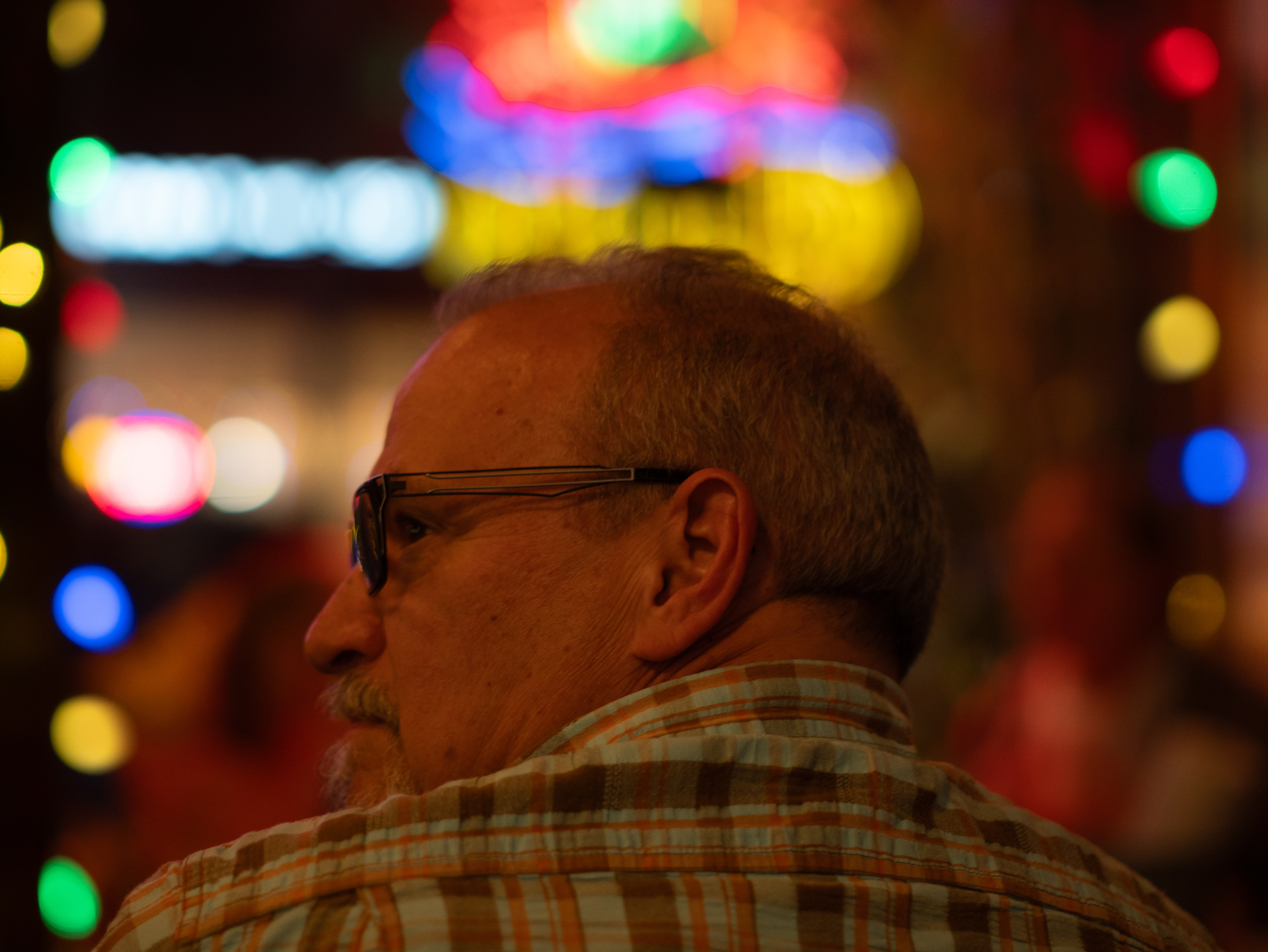 David Cuthbert, a middle-aged man with short, graying hair and glasses is seen in a dimly lit environment with colorful bokeh lights in the background. He is wearing a plaid shirt and appears to be looking off to the side with a contemplative expression. The vibrant, out-of-focus lights suggest a lively nighttime setting, possibly a bar or a festive outdoor space.